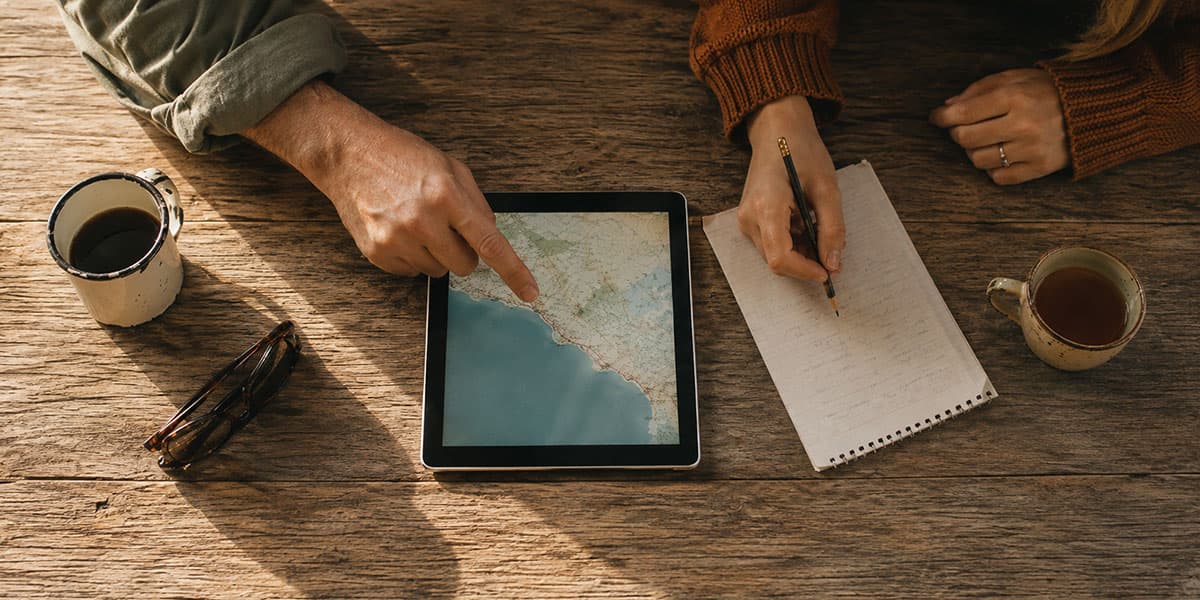 Hands tracing a coastal route on a tablet map, beside a notebook and coffee
